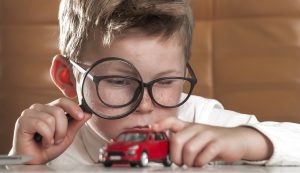 Child boy with magnifying glass inspecting red toy car. Concept of choice. Magnifier helps expert to estimate the benefits. How to choose transport